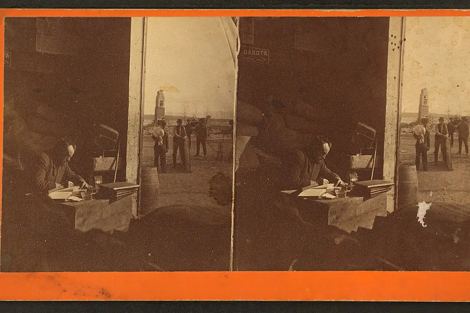 A man working at a desk in a barn or shed with the ruins of Kineo house, after a fire, in the background, from Robert N. Dennis collection of stereoscopic views